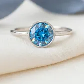 A ring with a bright blue round zircon in a beaded setting, on ivory linen