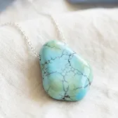 A freeform turquoise pendant with dark veins on a fine silver chain, on ivory linen